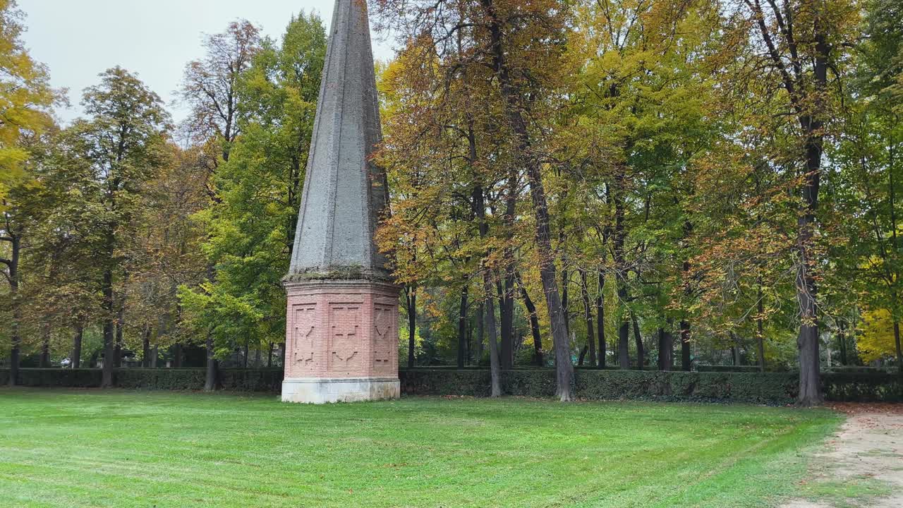 Imágenes a cámara lenta muestran la geometría detallada de un obelisco histórico en los Jardines Reales de Aranjuez, elevándose sobre césped recién cortado, enmarcado por árboles con los colores del otoño.