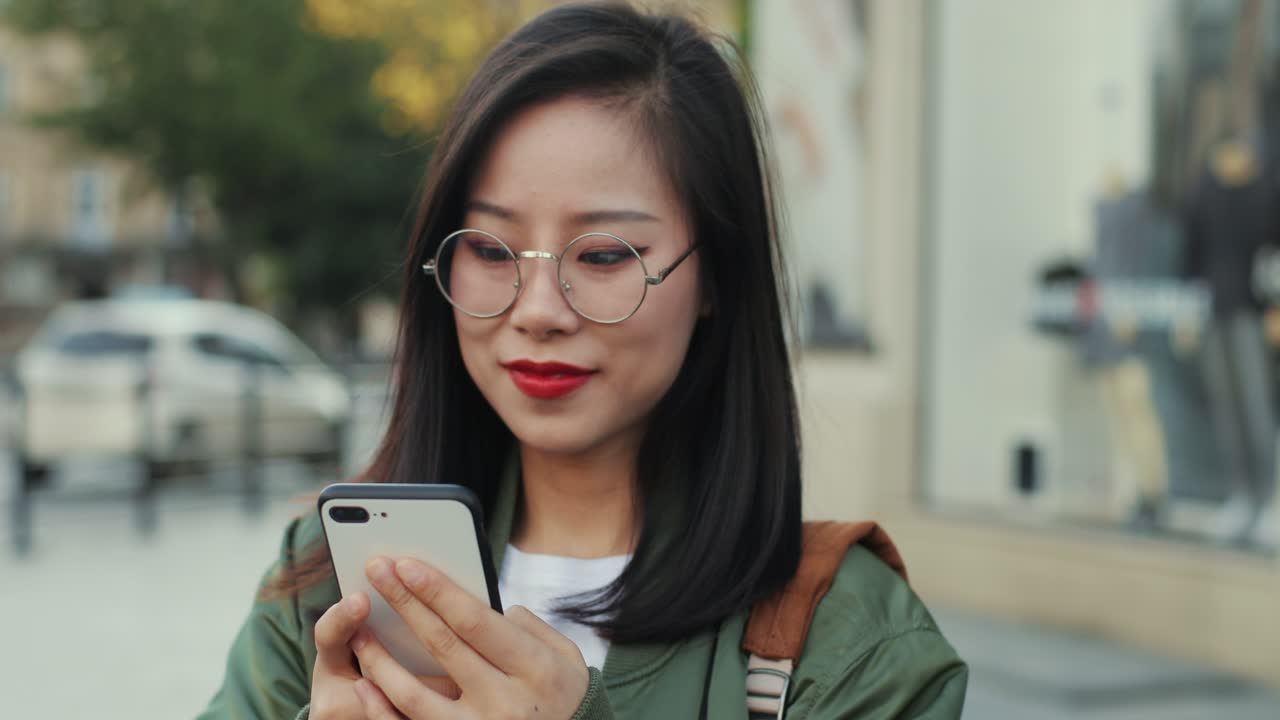 primer plano de la alegre joven con anteojos tocando y enviando un mensaje de texto en el dispositivo de teléfono inteligente en las manos