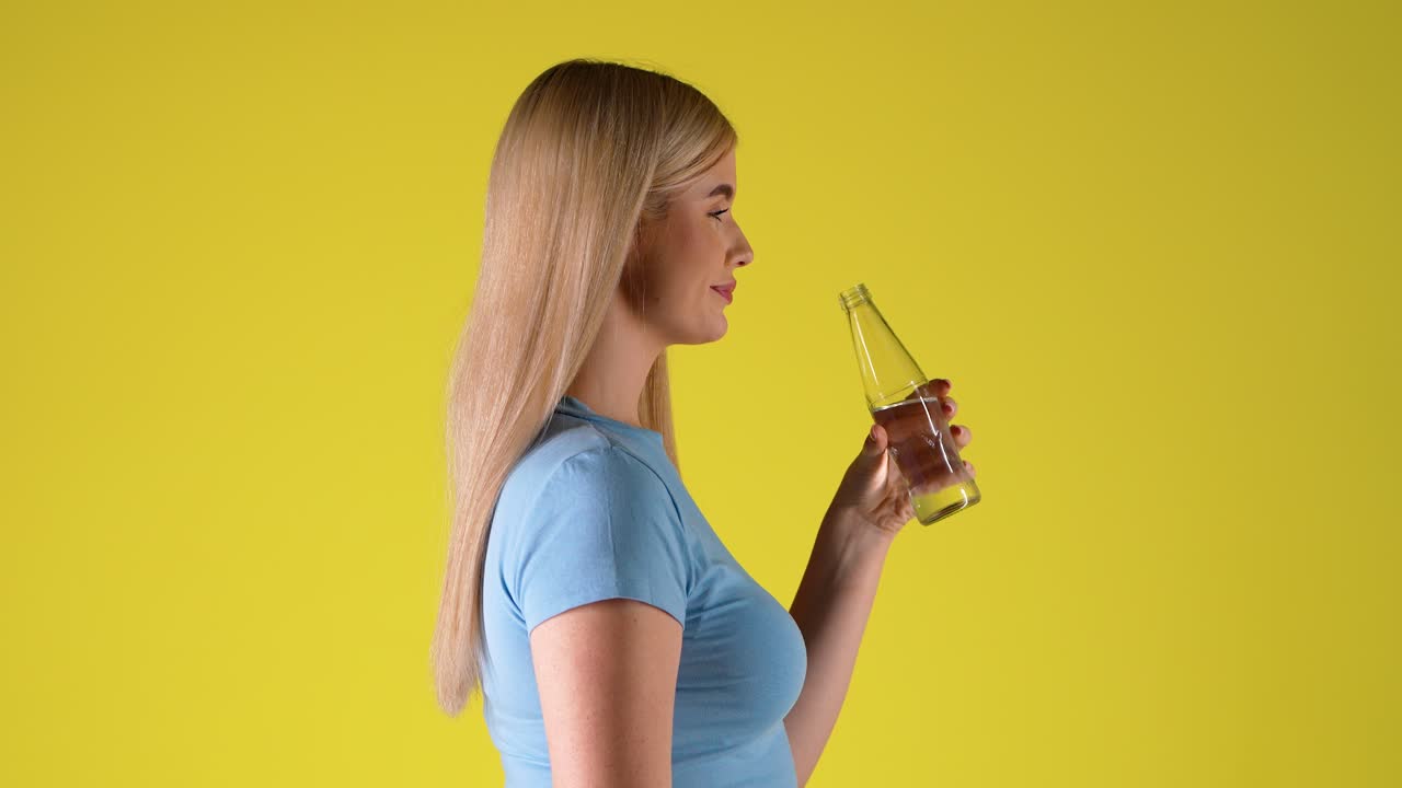 Side View Of Blonde Woman Drinking Water From Glass Bottle, Studio Portrait