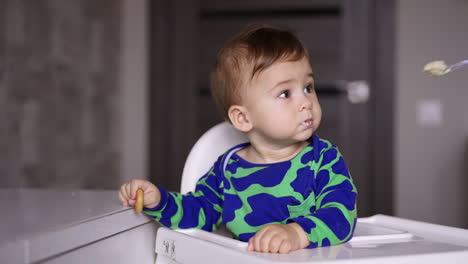 mom's hand shoves a full spoon with porridge to her child. cute baby turns away but then opens mouth. blurred backdrop indoors.