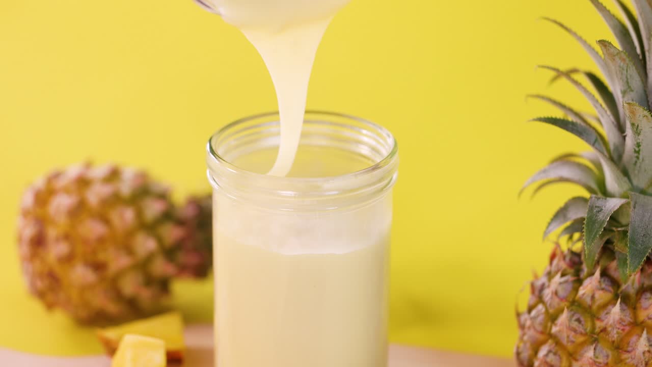 Pineapple smoothie pours into glass jar with whole fruit, cubes, bright lighting, vibrant yellow backdrop
