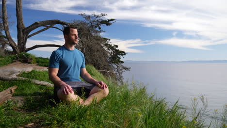 a fit young caucasian man sitting down to practice meditation in and mindfulness in nature on a cliff over the blue ocean and beach of santa barbara, california