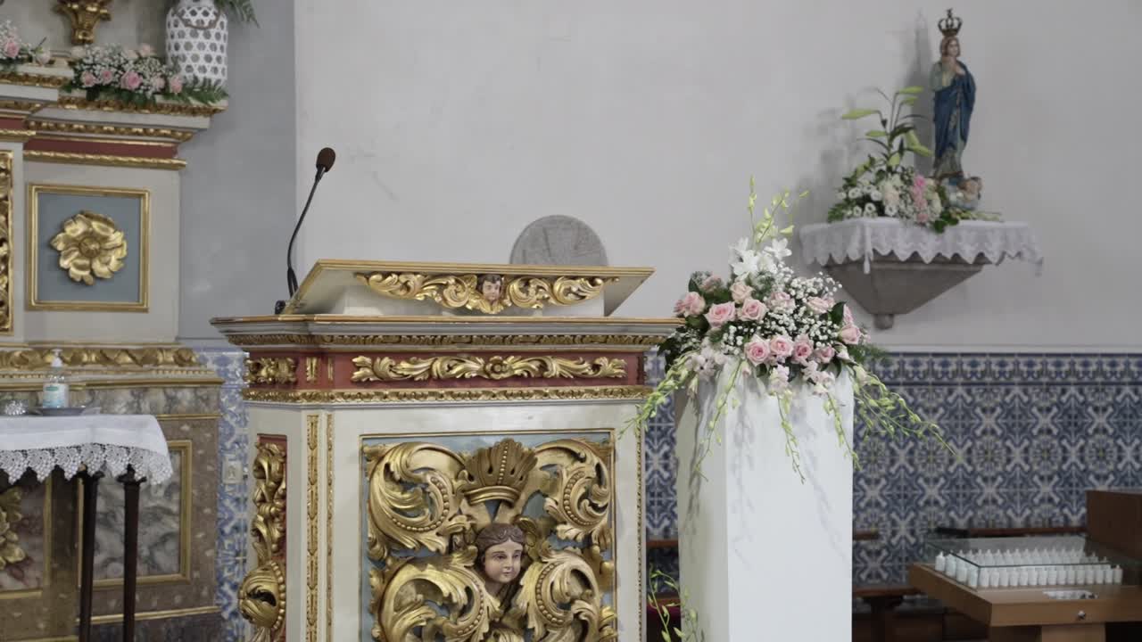 Pulpito di chiesa dorato con microfono, fiori, pareti piastrellate e una statua mariana.