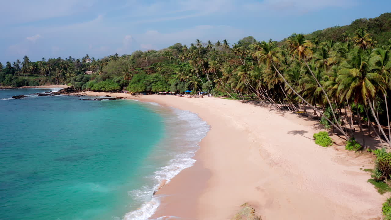 Slow aerial push-in shot over a sandy tropical beach with palm trees and straw umbrellas near coastal villas at Silent Beach, Sri Lanka during midday.