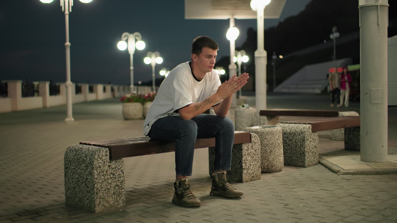 hombre sentado en un banco al aire libre por la noche con las manos cubriendo su cara y quitándola, pareciendo estresado o ansioso, las luces de la ciudad y los peatones lejanos crean una atmósfera urbana tranquila en el fondo