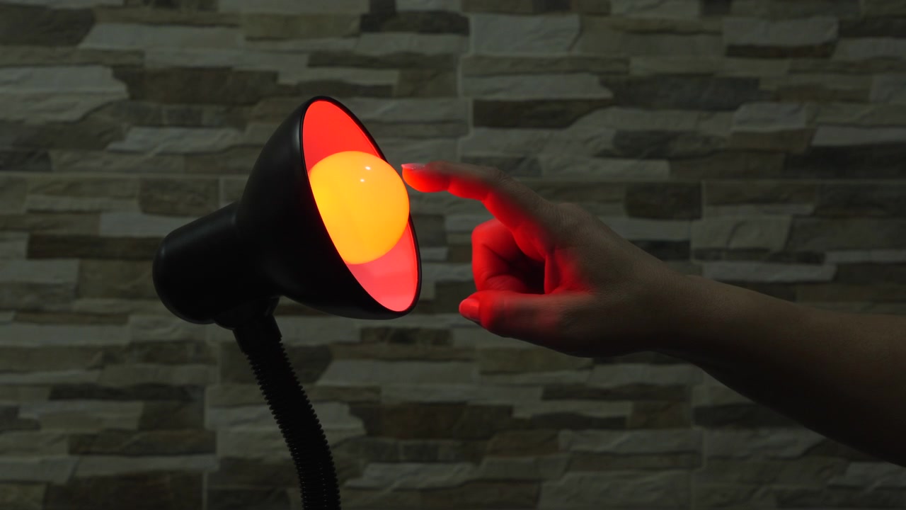 En hand sträcker sig in för att tända en liten skrivbordslampa, som övergår från mörker till ett varmt rött sken mot en vägg med stenstruktur.