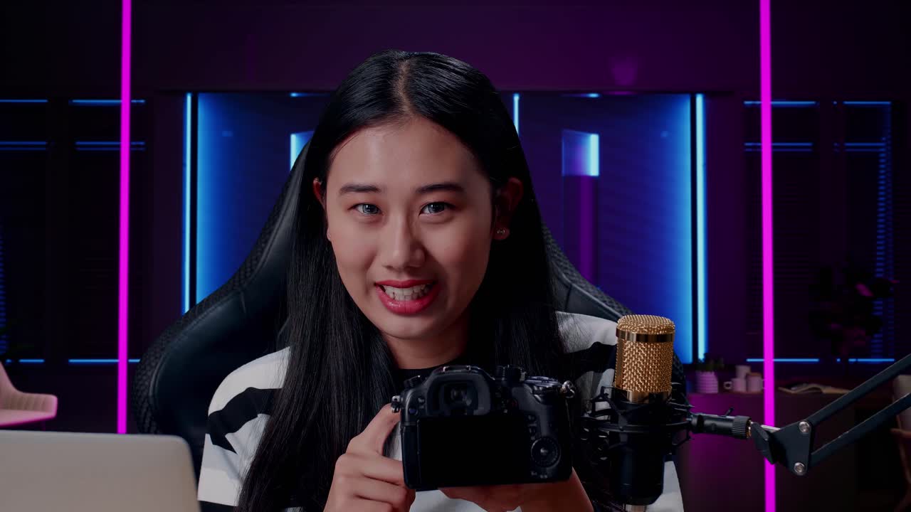 컴퓨터로 미소 짓는 아시아 여성의 클로즈업 카메라가 레트로에서 네온 조명으로 조명된 방에서 검토됩니다.