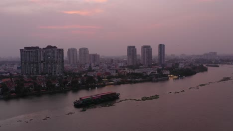 Container-boat-carrying-cargo-on-Saigon-River,-Vietnam-at-sunset-with-drone-view-of-water-and-city-skyline