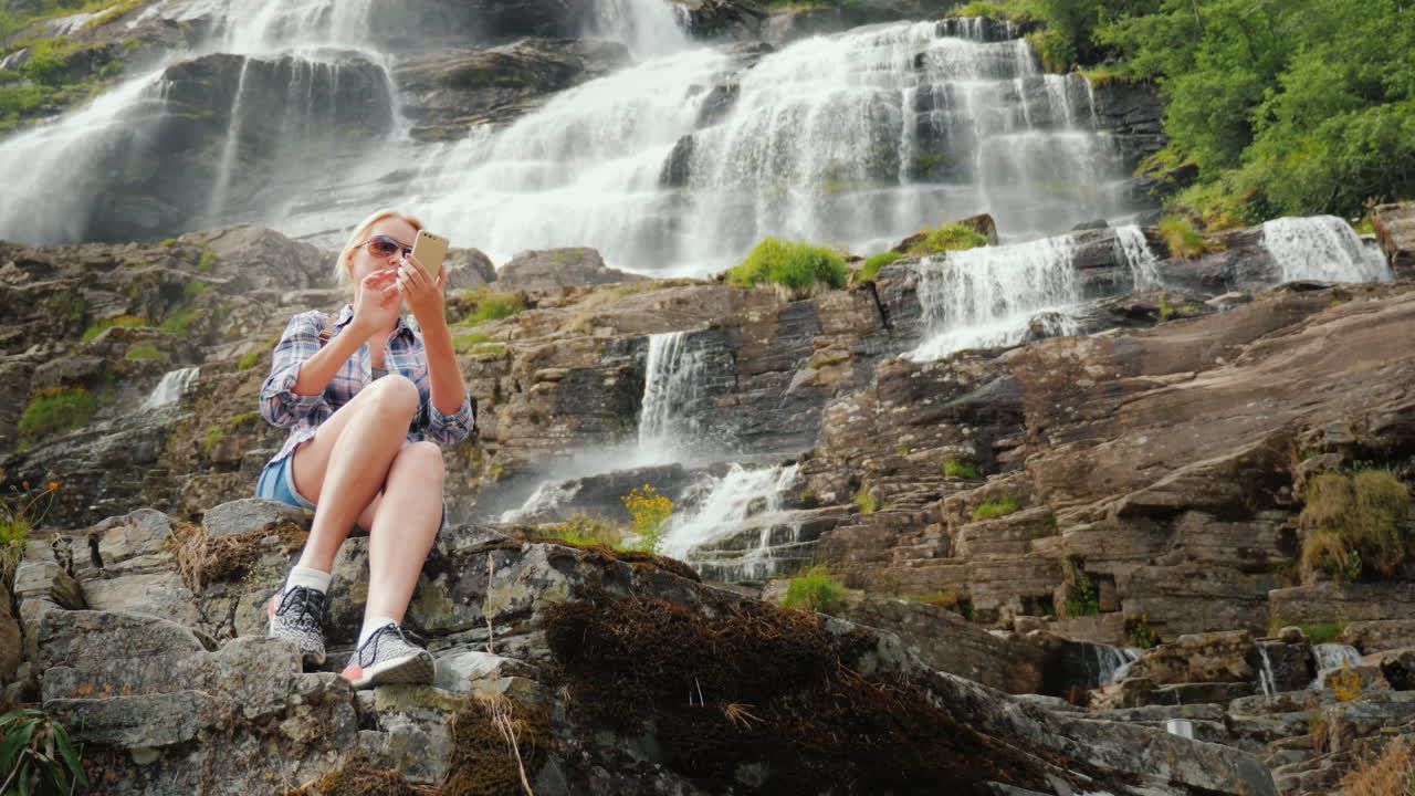un viajero usa un teléfono inteligente contra el fondo de una cascada tvindefossen en noruega 4k video
