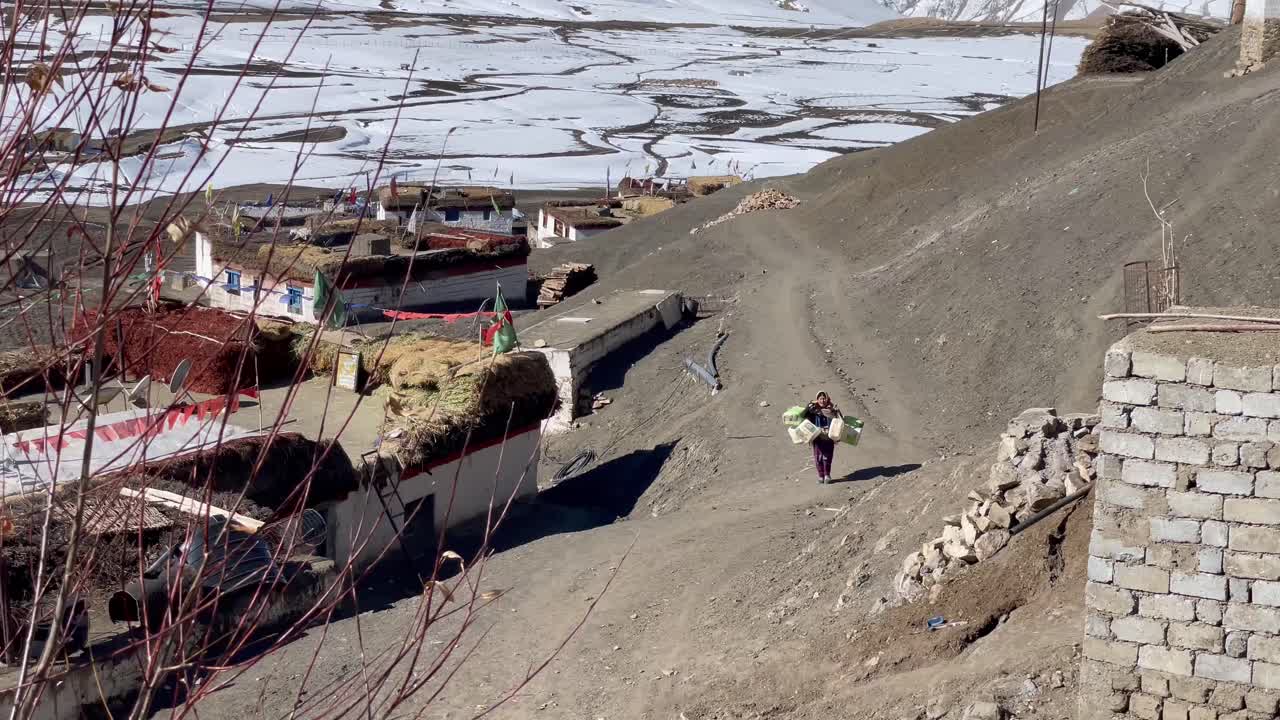 Un abitante del villaggio locale che trasporta un carico di contenitori di plastica camminando su un sentiero brullo e polveroso in salita nella Valle dello Spiti, Himachal Pradesh, India