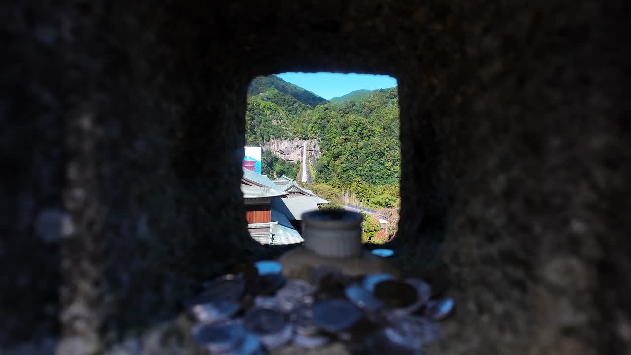Scenic view of Nachi Waterfall framed by a stone lantern with coins offered in the foreground, capturing a blend of nature and tradition