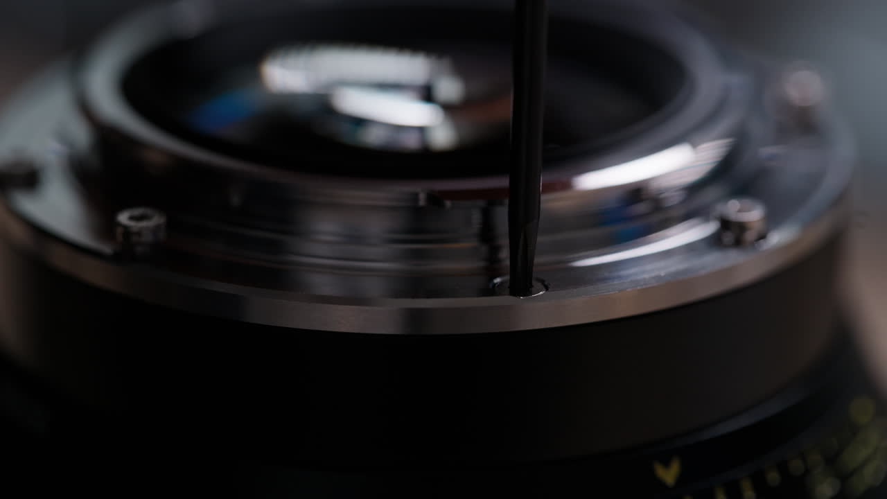 Low angle side view of hands removing a Torx screw from a cinema lens mount during EF to PL conversion. Screwdriver and lens body clearly visible. Detailed technical modification footage