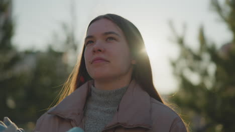 un primer plano de una mujer joven con una chaqueta de melocotón y un suéter de cuello gordo gris, de pie en un parque iluminado por el sol. ella está sosteniendo un par de patines y mirando hacia arriba con una expresión reflexiva