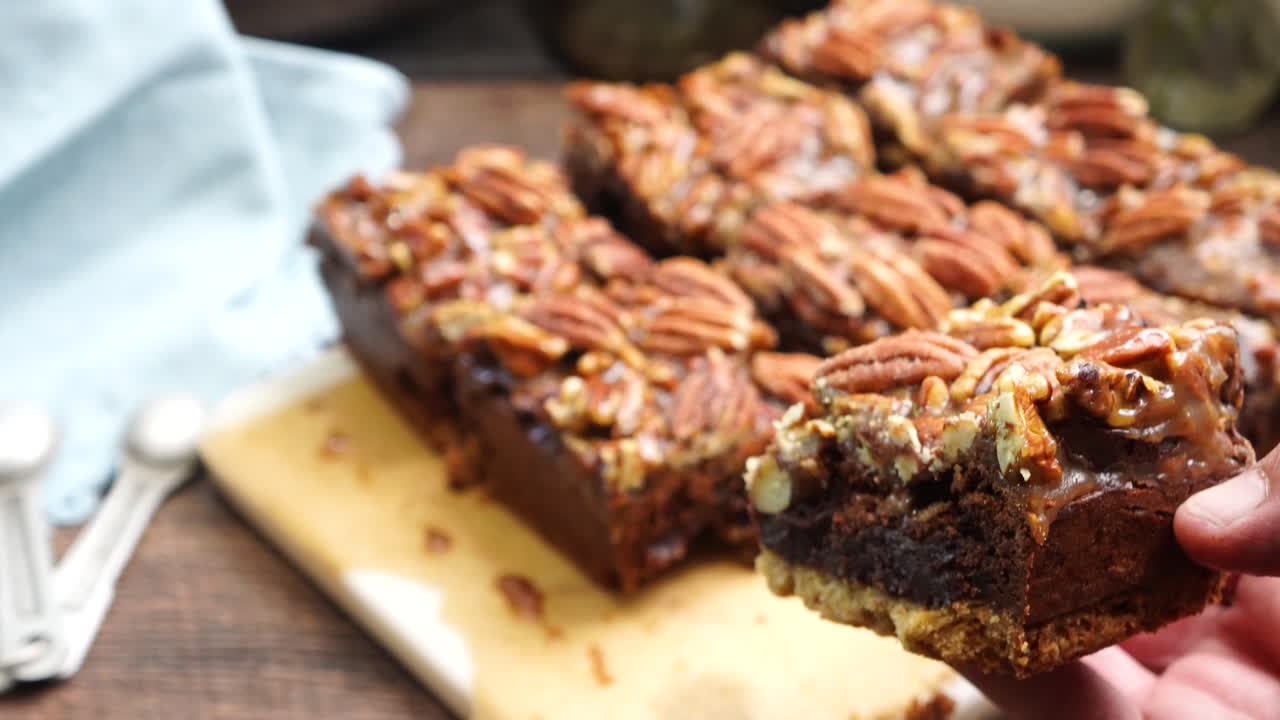 Baking delicious caramel pecan brownies. Using hands to pick up brownie showing a close up. Freshly baked chocolate brownies with sweet caramel and nutty pecans. Brownies are on a cutting board.