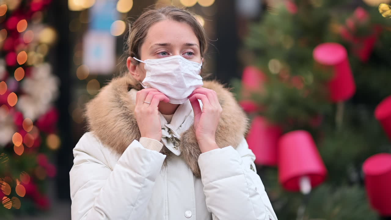 A woman in a white coat prepares to put on a face mask while surrounded by colorful holiday lights and decorations in a busy city area. The atmosphere is vibrant and cheerful