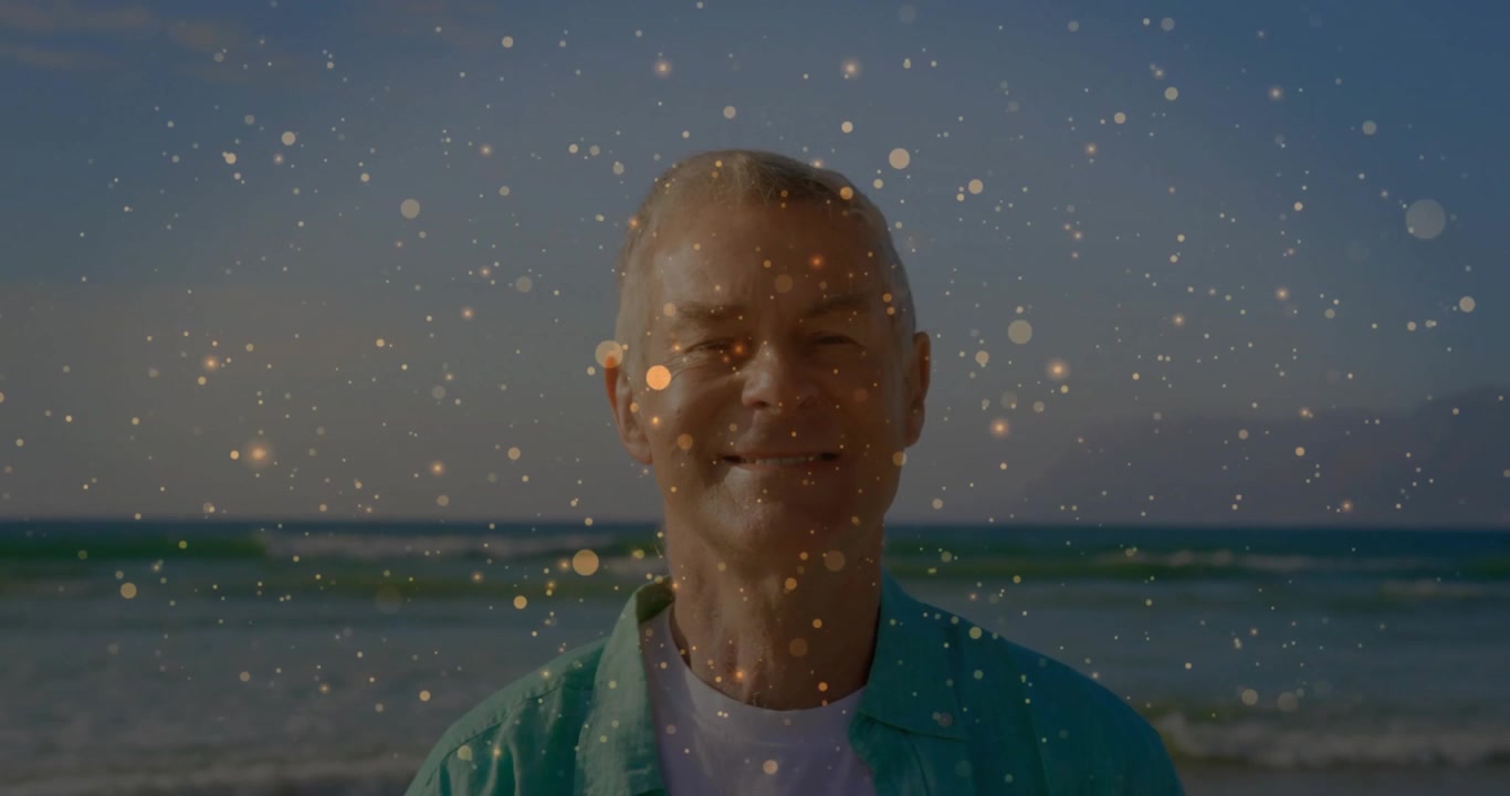Senior man pausing on ocean beach, golden particles drifting, intensifying then fading in wellness