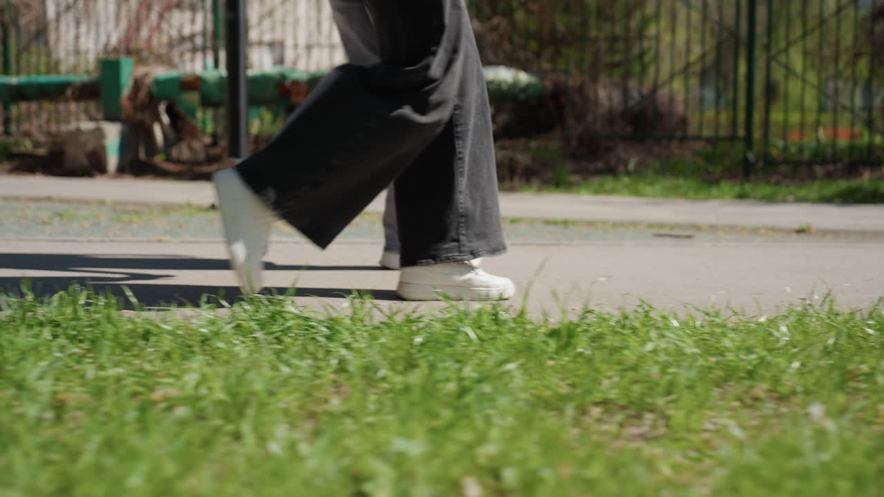 풀밭이 보이는 클로즈업 스니커즈가 길 위를 걷고 있어요. 흰색 밑창과 끈, 흔들리는 통 넓은 바지, 도시 울타리 배경, 일정한 측면 트래킹 숏으로 발의 움직임을 자세히 보여줘요.