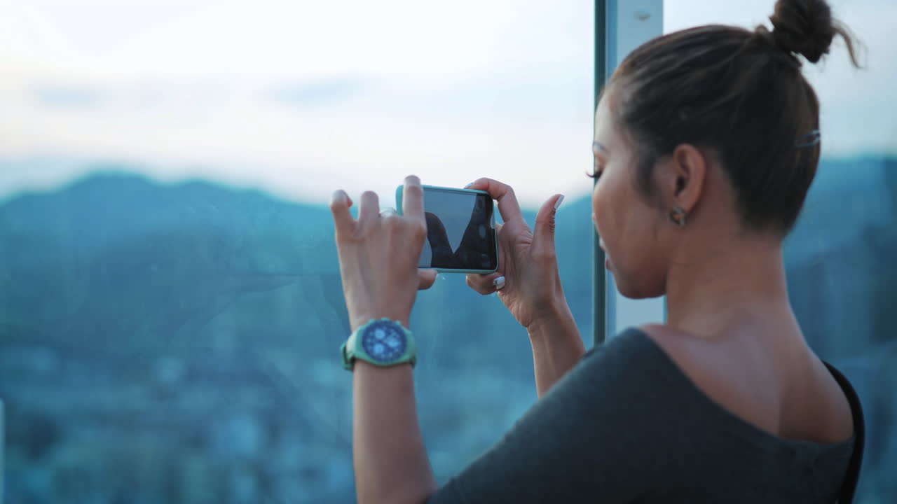 mujer con el cabello marrón claro atado en un pan, tomando una foto de la ciudad a través de una ventana, captura su enfocado en la tarea, vista del entorno urbano en el fondo, destacando la exploración de la ciudad