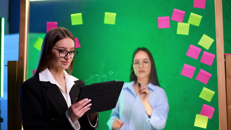 woman in glasses, black jacket and white shirt holds a tablet. brunette lady writes numbers on the glass wall.