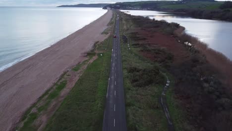 Aerial-footage-following-a-road-in-South-Devon-with-some-cars-on,-with-a-beach-on-one-side-and-lake-on-the-other