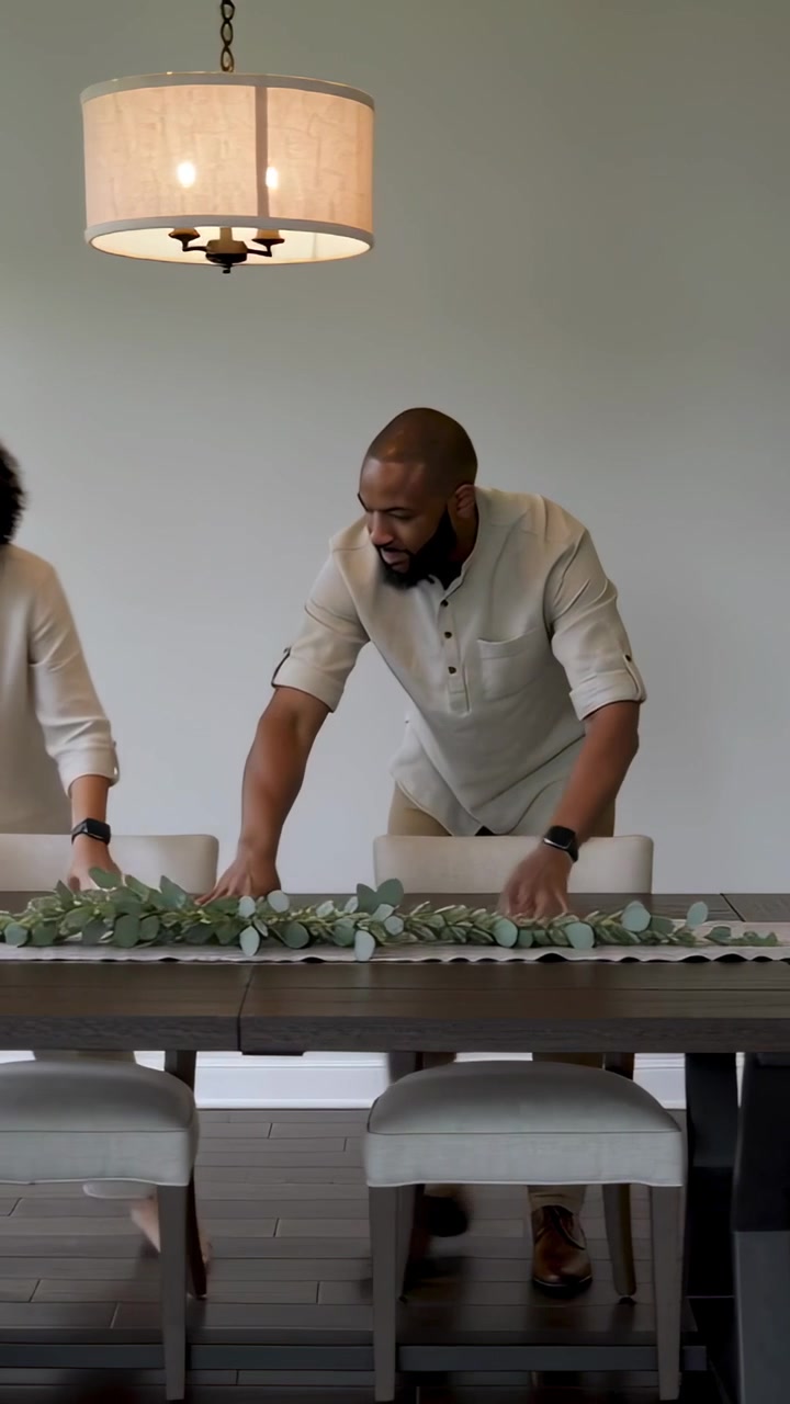 Vertical video: Arranging family aligning garland on dining table in linen shirts preparing dinner