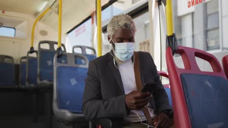 hombre mayor afroamericano con máscara facial usando un teléfono inteligente mientras está sentado en el autobús