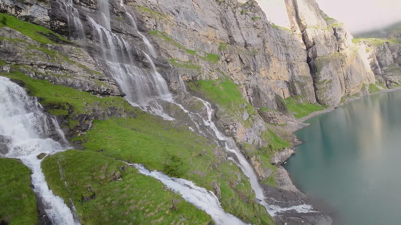 vuelo aéreo junto a una hermosa gran cascada en un paisaje montañoso, avión no tripulado volando sobre un lago azul - lago oeschinen, suiza