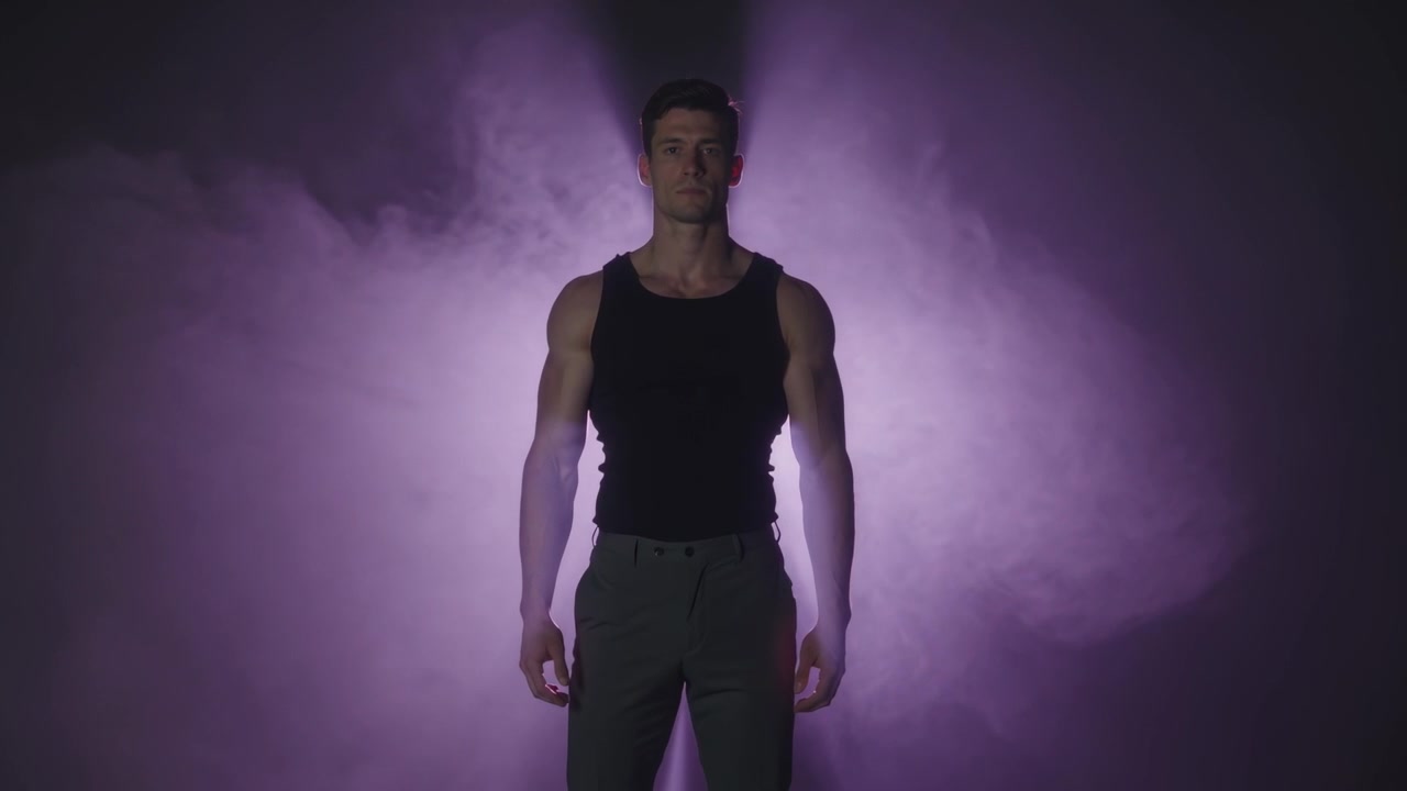 Controluce crescente, uomo con canotta nera e pantaloni grigi che mantiene una posa in studio, foschia che aggiunge luce di contorno.