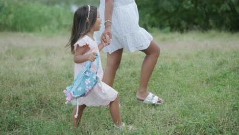 a pregnant woman and her young daughter, both dressed in white, walk hand-in-hand through a grassy park. the scene captures a serene moment of family bonding in nature, surrounded by trees.