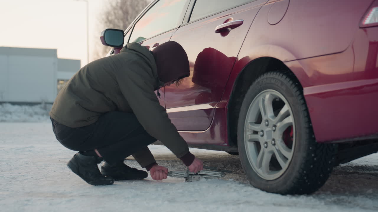 Young man squats next to red car in winter setting, placing metal jack under vehicle and preparing to loosen bolt on front wheel, surrounded by snow and soft morning light reflecting on car body