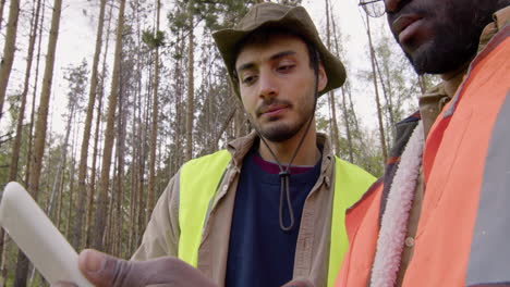 Caucasian-man-activist-and-african-american-man-watching-something-in-a-tablet-in-the-forest-while-they-deciding-where-to-plant-trees