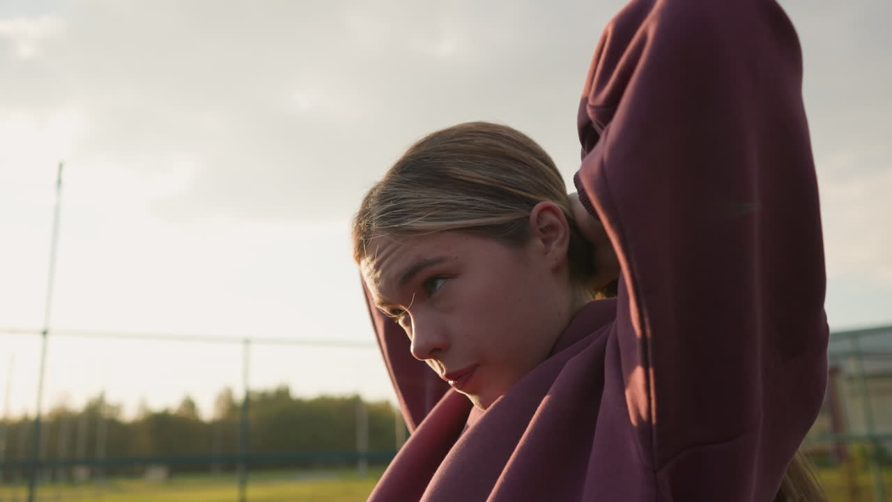 боковой вид женщины, связывающей волосы в обратном направлении на открытом воздухе, солнечный свет создает светящийся эффект вокруг ее руки, мягкий солнечный луч подчеркивает ее черты, когда она готовится к активным действиям на свежем воздухе в естественной обстановке