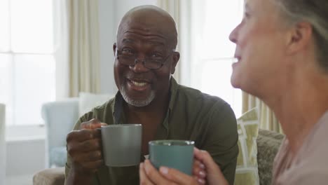 Glückliches-älteres,-Vielfältiges-Paar,-Das-Hemden-Trägt-Und-Kaffee-Im-Wohnzimmer-Trinkt