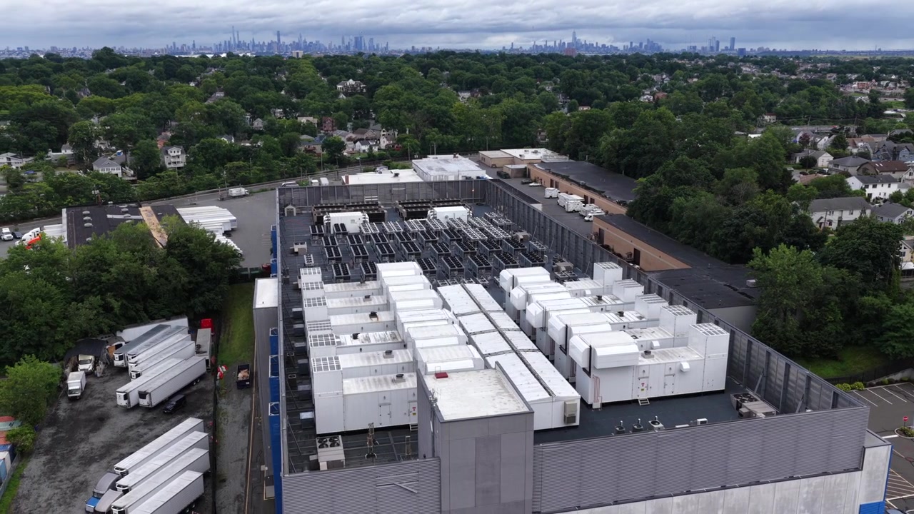 Rækker af store hvide kølegeneratorer fylder et datacenter-tag i Clifton, New Jersey, fjerne tågede Manhattan skyline rejser sig langt ud over tætte grønne forstadstage. Luftfoto drone-orbit vidvinkeloptagelse