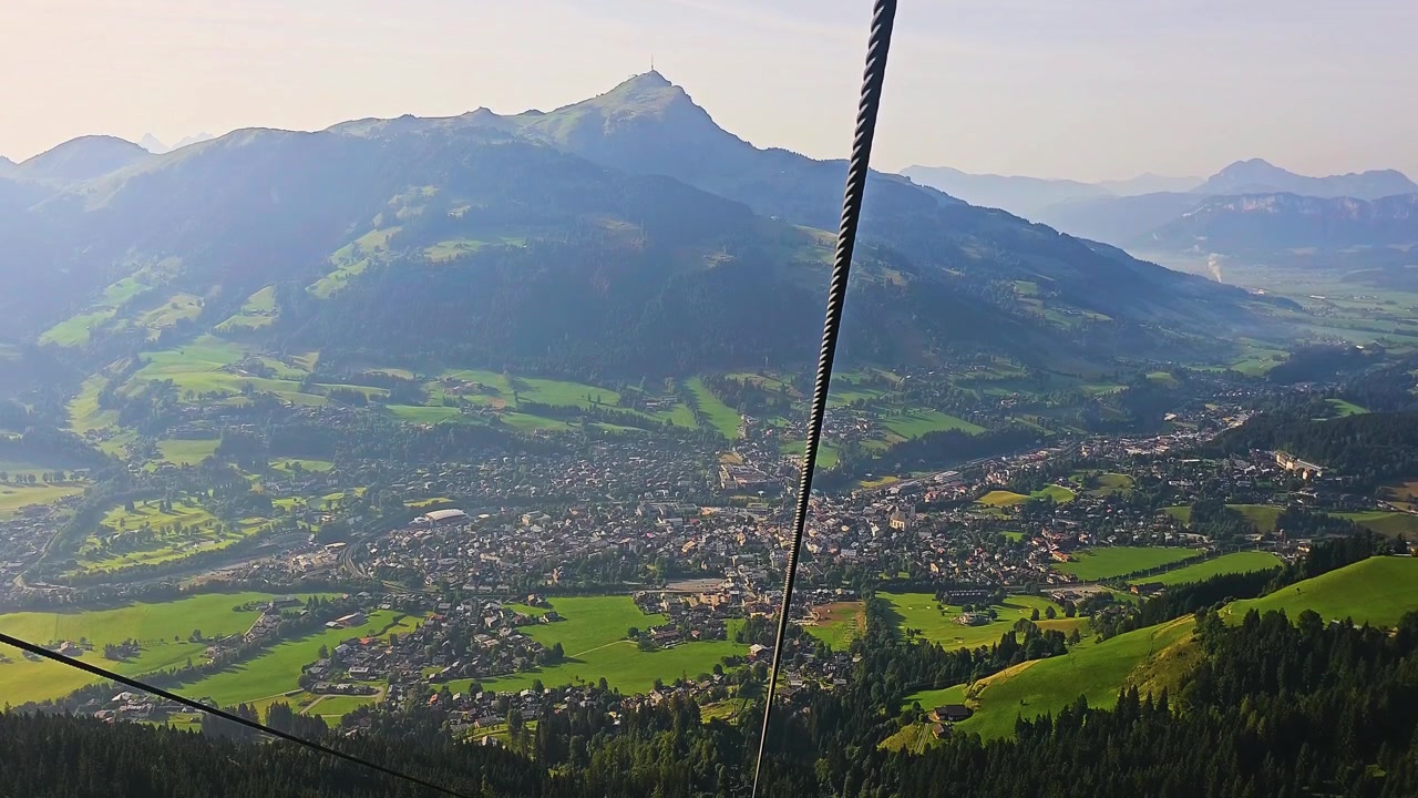 Ampia vista aerea da una funivia di montagna, con cavi che attraversano l'inquadratura, valle alpina verde, tetti delle case, pendii boscosi e cime montuose spettacolari in una calda luce estiva