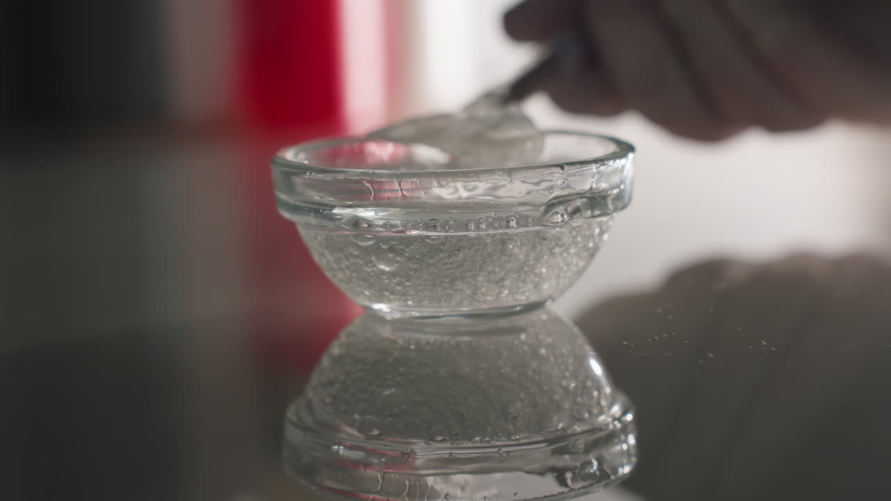 Face mask gel bubbles shimmering on glass surface with expert gloved hand dipping brush into small bowl creating reflective skincare prep scene under soft ambient light for beauty treatment setup