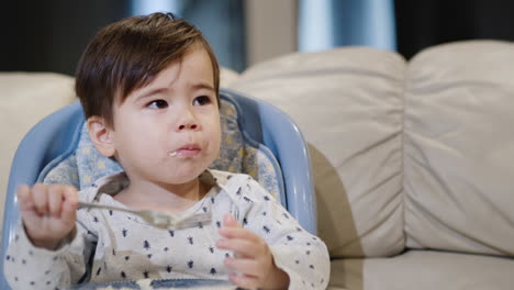 cute asian baby independently eats porridge from his plate, sits in a chair for feeding