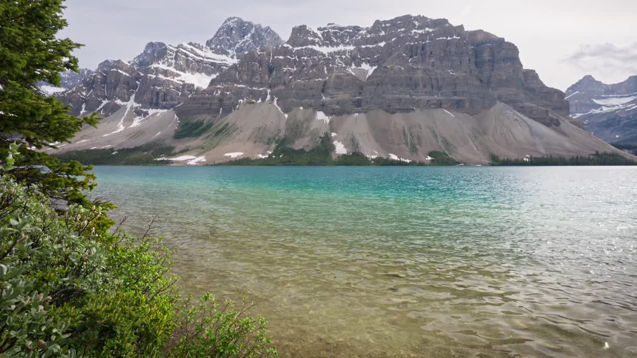 La camera esegue una panoramica verso l'alto per rivelare lo splendido lago Bow color turchese, alimentato dai ghiacciai, in Alberta, Canada, nel Banff National Park, con onde che lambiscono delicatamente la riva di fronte a enormi montagne scoscese