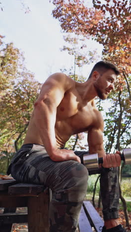 shirtless sportsman doing workout in autumn park. active man with muscular body sitting on a bench and lifting dumbbell on nature background. vertical video
