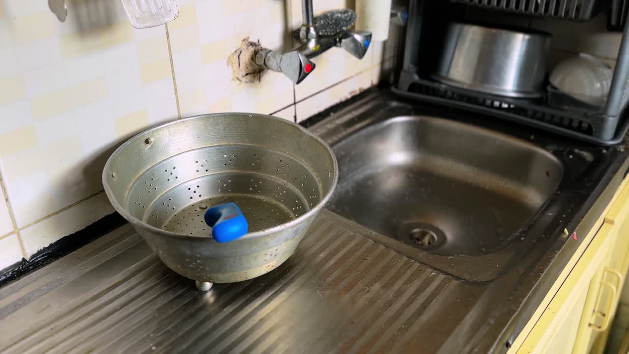 Gammel metaldørslag med en blå rengøringssvamp placeret på en drænbakke af rustfrit stål ved siden af en køkkenvask med væghane og flisebelagt baggrund i naturligt indendørs lys.