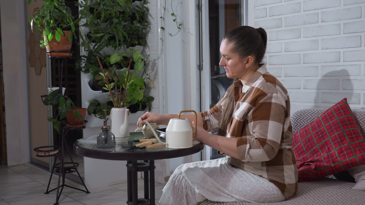 Young woman with focused expression seated on sofa beside indoor greenery sprinkles seeds into black plastic tray on glass table surrounded by watering can, small gardening tools, cozy home setting