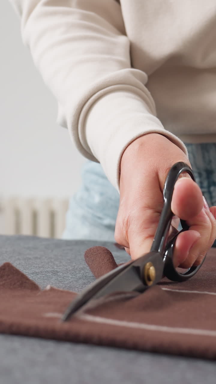 Closeup hands cutting brown wool fabric on table. Tailor marking chalk pattern, cutter slicing sleeve, sample maker trimming seam in sunlit studio denim jeans and neutral sweater visible precise