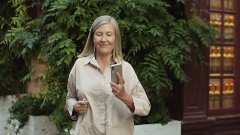 Senior-Happy-And-Cheerful-Woman-With-Short-Gray-Hair-Listening-To-Music-Via-Headphones-On-Smartphone