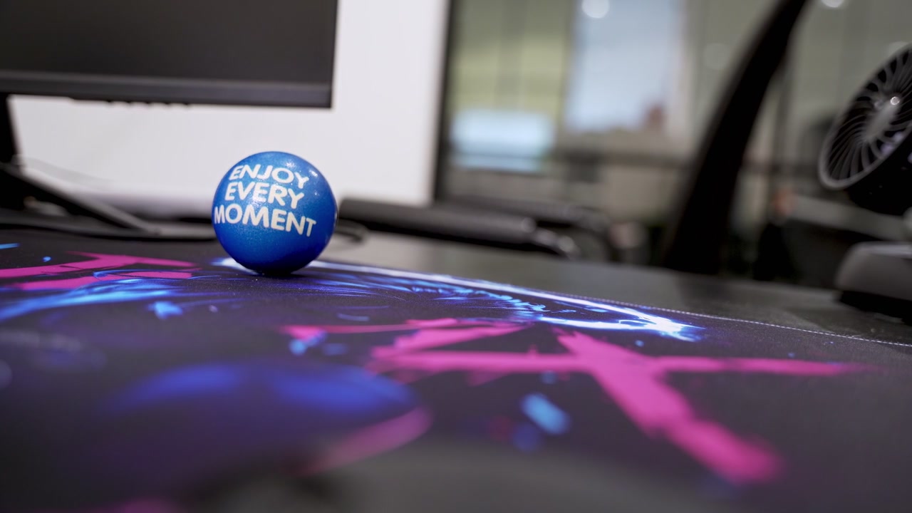 Close-up of a hand squeezing a blue stress ball reading "Enjoy Every Moment" on a colorful mousepad at an office desk. A computer monitor and blurred modern office environment are in the background.