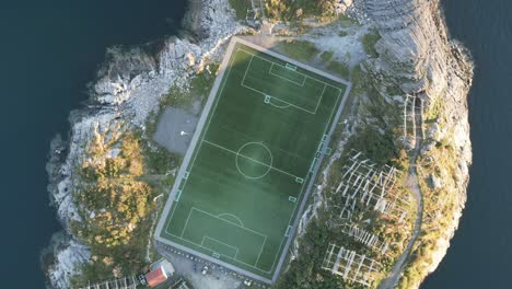 rotating upwards aerial footage showing the football soccer stadium in henningsvaer, lofoten, norway with its classic view on an island right by the ocean and surrounded by tall mountains