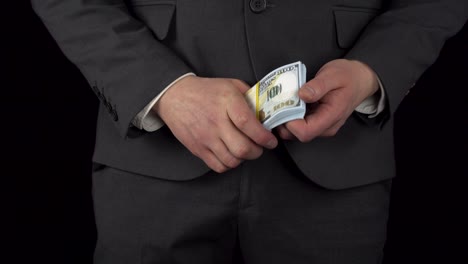 a young businessman in a suit takes out a pack of dollars from his pocket and holds it in his hands. the waist of a man close-up on a black background.