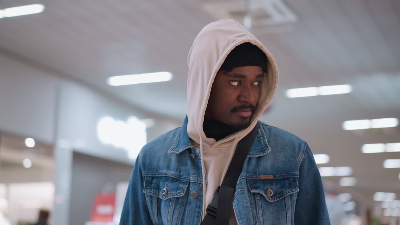 young black man wearing hoodie and denim jacket walking inside shopping mall holding green shopping bag at side gazing forward through store window display, casual urban retail lifestyle scene