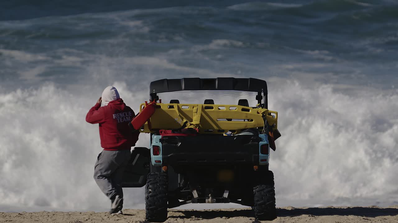 llifeguard en una sudadera con capucha roja sale de un duny buggy en la playa frente a enormes olas en el fondo