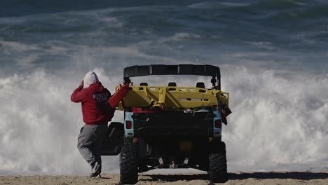 llifeguard in a red hoodie gets out of a duny buggy on the beach in front of huge waves in the background