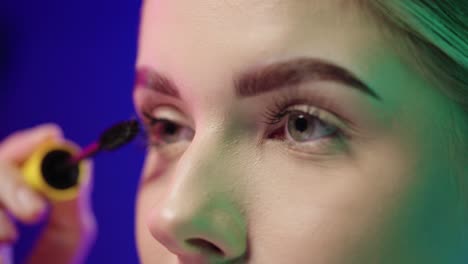 close up of the eyes of a young beautiful woman while she is fixing her eyelashes with a mascara before going out or partying in the club in front of blue background in slow motion